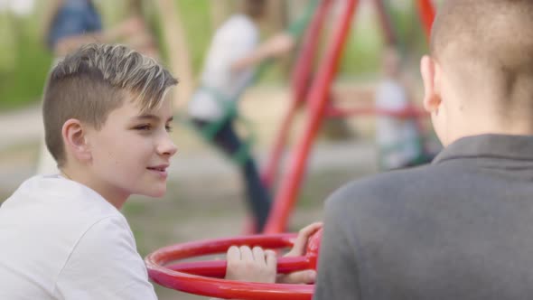 Two Brothers Sitting in the Park and Talk. Woman Swinging Boys at the Background