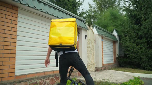 Tracking Shot of Caucasian Courier Riding Bike Walking to Fence Gate Pressing Door Bell