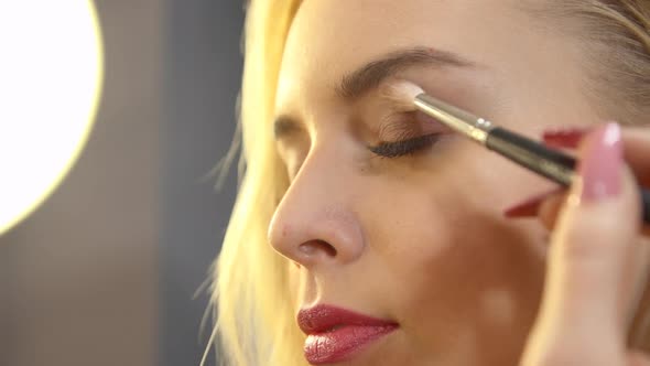 Young Stylist Putting Makeup on Face of Beautiful Blonde Girl