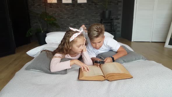 Children - a boy and a girl read a thick book with a magnifying glass, lying on the bed at home