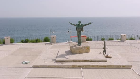 Slow aerial flyover of a statue of a man with his hands raised in Polignano a Mare, Italy.