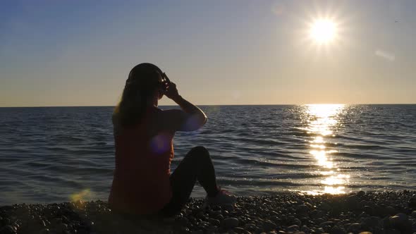 Girl listens to music wearing headphones on seashore at sunset
