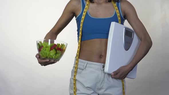 Woman in Loose Pants Standing with Scales, Holding out Bowl of Salad, Dieting