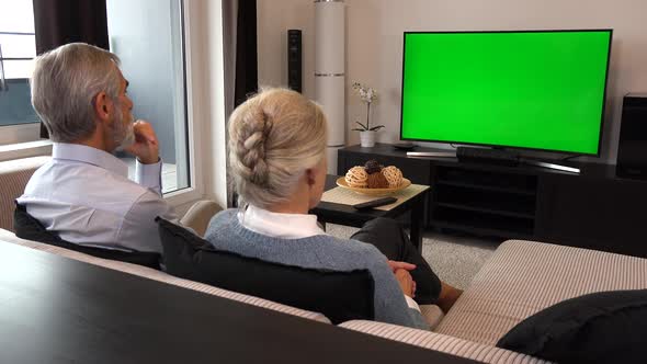 An Elderly Couple Sits in A Living Room in An Apartment and Watches TV with A Green Screen