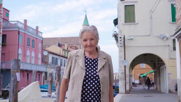 Old Woman Stands on Channel Embankment Street with Houses