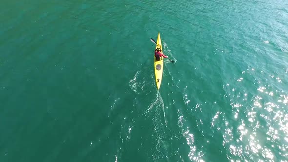 A kayaker paddles in a scenic mountain lake with a drone hovering above him.