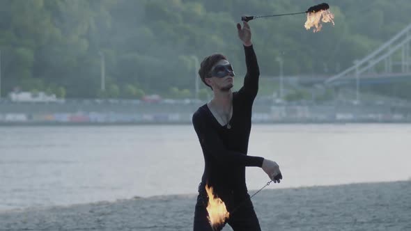 Young Slim Man in Black Clothes and Mask Performing a Show with Flame Standing on Riverbank