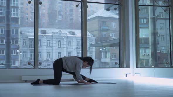 Young Woman Stretching Legs on Mat in the Dancing Hall with Large Floor-to-ceiling Windows