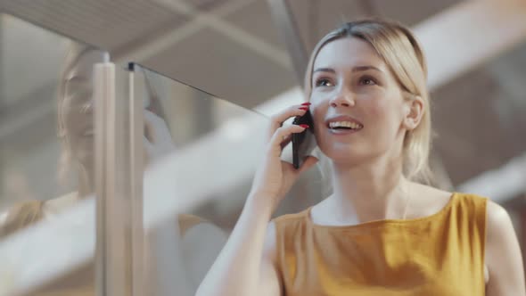 Young Blonde Businesswoman Leaning on Glass Wall and Chatting on Phone