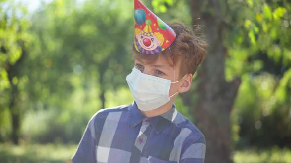 Sad Caucasian Teenage Redhead Boy in Coronavirus Face Mask and Party Hat in Sunrays Outdoors Alone