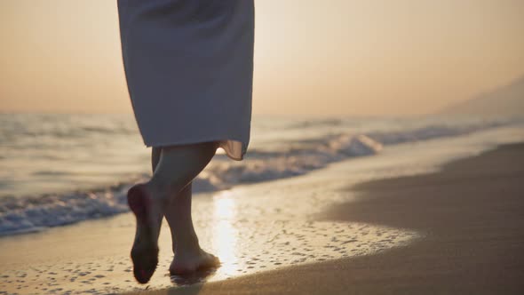 Bare feet of a young girl in a white dress walking along the shore on a summer evening
