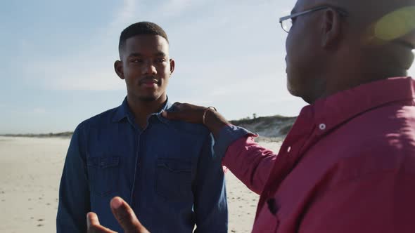 African american father standing on beach with teenage son, putting hand on his shoulder and talking
