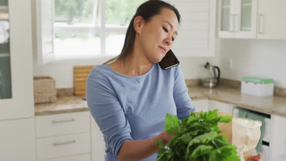 Happy asian woman unpacking groceries and using smartphone in kitchen