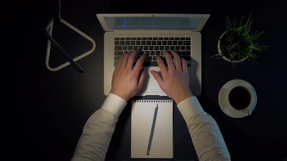 Male Businessman in White Shirt Sitting at the Table in the Evening Working on a Laptop and Drinking