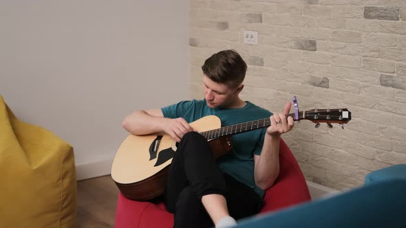 Young Man Is Playing at the Guitar Sitting on a Red Bean Bag in a Living Room