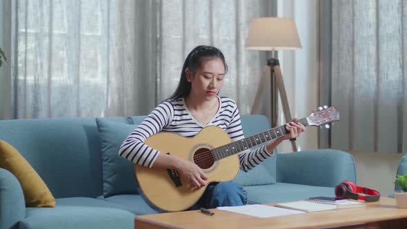 Asian Woman Composer Looking At Paper On Table And Playing A Guitar At Home