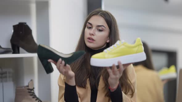 Thoughtful Millennial Woman Holding Casual Yellow Sneakers and Elegant Green Highheels Thinking