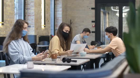 Young Caucasian Men and Women in Covid Face Masks Sitting in Cafe Surfing Internet