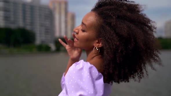 Portrait of a Young African Woman in a Lilac Top with Ties and Puffy Sleeves on the City Embankment