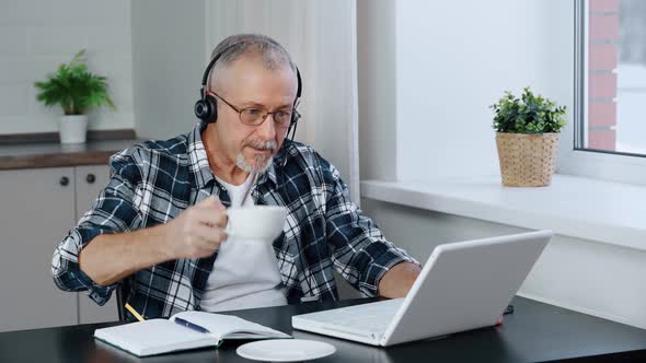 An Elderly Businessman Wears Headphones Talks on a Conference Call While Working