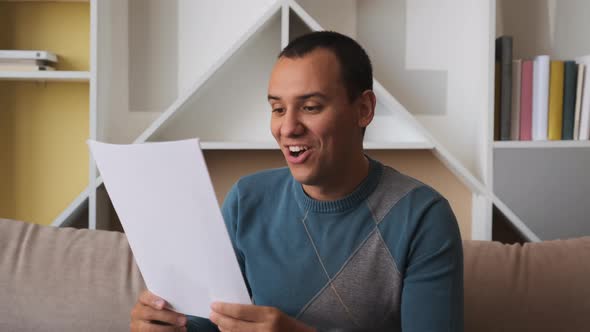 Happy Young Man Reading Paper Letter, Amazed By Good News, Sitting on Sofa at Home
