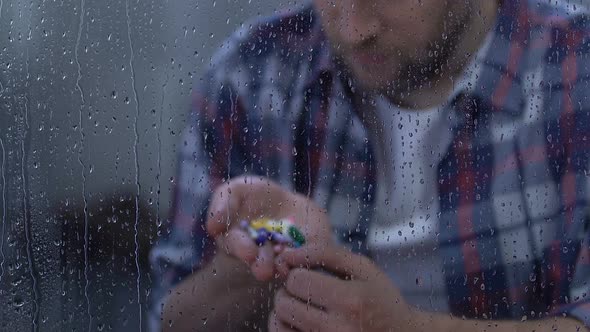 Depressed Sick Man Holding Full Palm of Medication, Sitting Behind Rainy Window