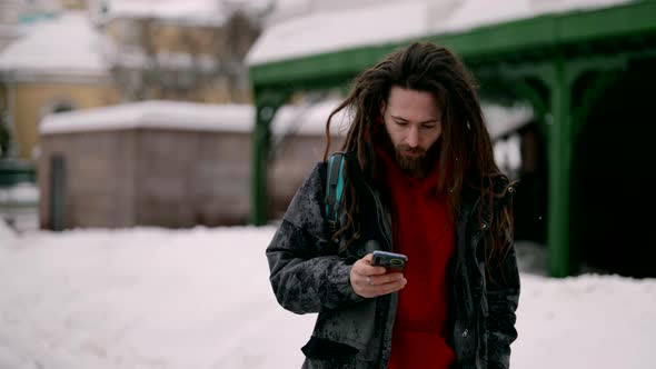 Young Stylish Man with Dreadlocks is Reading News in Smartphone on Street in Winter