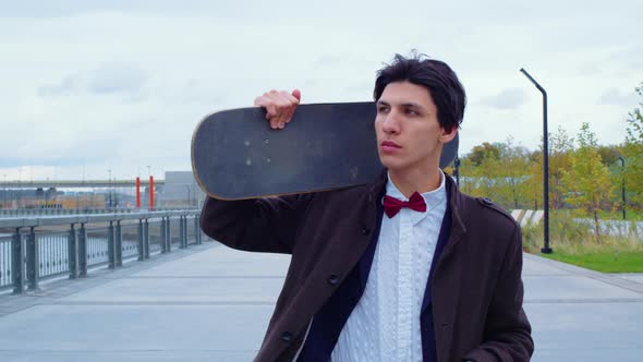 Boy in Coat with Skateboard Walks on Embankment