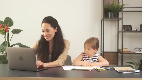 Young Mother Smiling and Working with a Laptop at Home While Her Little Daughter Draws with Pencils