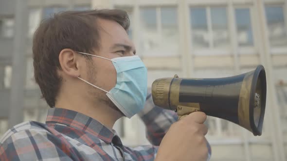 Side View of Young Male Activist in Face Mask Shouting Through Megaphone. Close-up Portrait