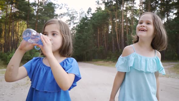 Two Cute Girls Are Walking Along the Forest Road and Drinking Water