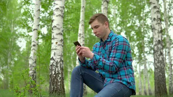 Young Man in Nature with a Phone in His Hands. A Man Sits on a Stump in a Birch Forest and Leads