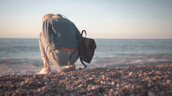 a blonde in a denim suit sits on rock on seashore against backdrop of sea sunset