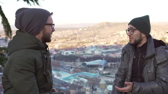 Two Caucasian Males in Hats and Glasses Sitting Opposite Each Other and Shaking Hands Outdoors in