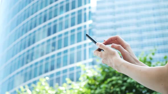 Women's Hands Use a Smartphone on the Background of a Glass Business Center and Green Trees