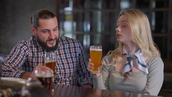 Positive Caucasian Couple Talking Clinking Glasses Toasting Drinking Beer Sitting in Pub Indoors