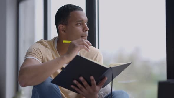 Portrait of Handsome African American Young Man Sitting on Windowsill Thinking and Writing in