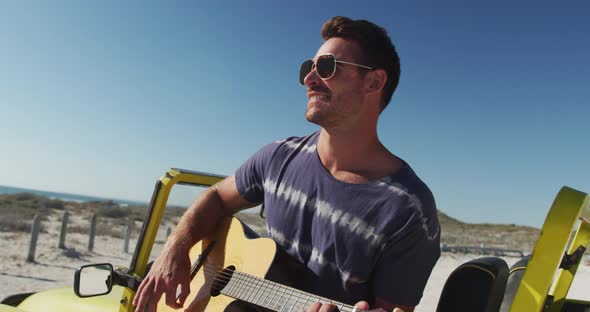 Happy caucasian man sitting in beach buggy by the sea playin gguitar