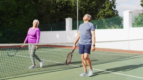 Video of happy biracial senior couple walking with rackets on tennis court