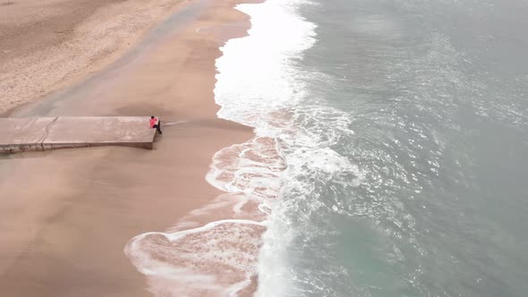 Lonely woman is relaxing by stormy sea sitting on edge of pier and swinging feet near water