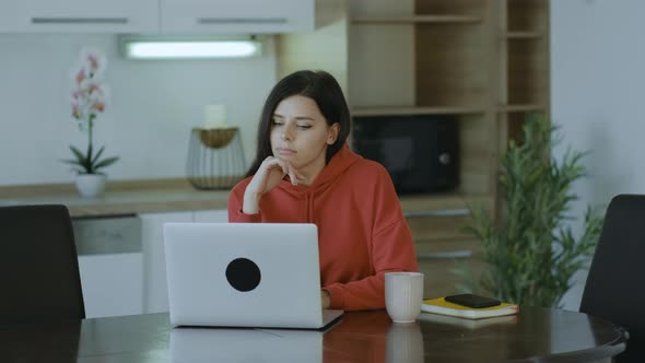 Sadness brunette woman in red hoodie  working online on laptop sit at home