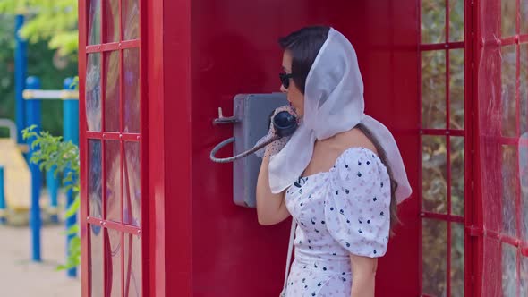 Beautiful Young Woman Happily Speaks on the Phone in an English Style Red Telephone Booth