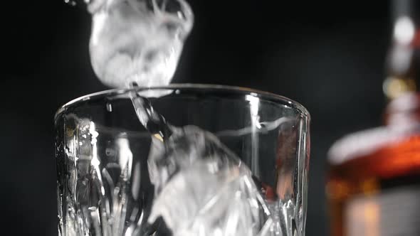 Ice Cubes are Being Dropped to the Glass in Slow Motion with the Bottle of Alcohol Drink in the