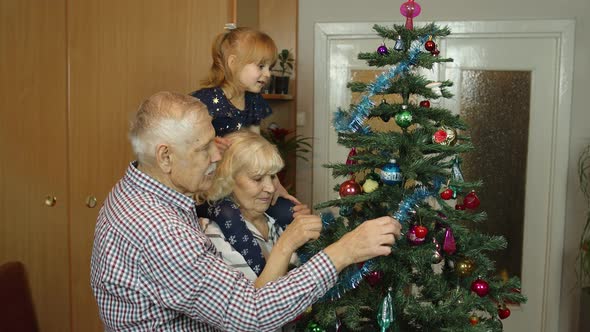 Children Girl with Grandparents Couple Decorating Artificial Christmas Pine Tree at Oldfashion Home