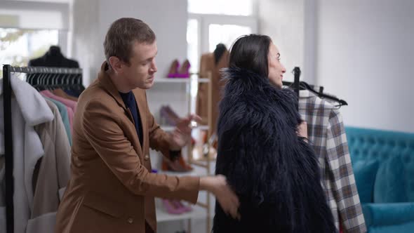 Caucasian Gay Man Helping Woman Choosing Fur Coat in Shop Indoors