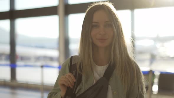 Portrait of Smiling Young Blonde Girl Traveller with Backpack Stands in Airport Terminal