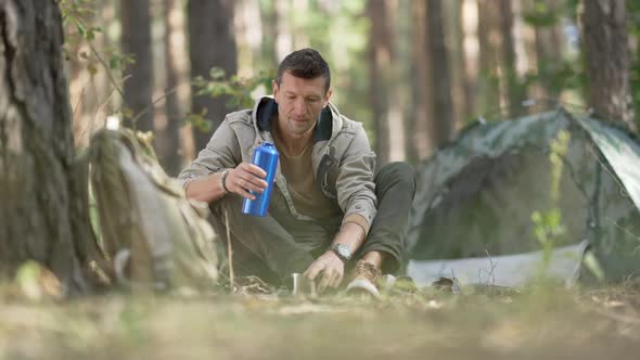 Front View Happy Male Tourist Pouring Drink From Thermos in Slow Motion Sitting at Campsite in
