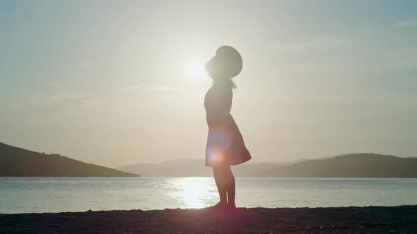 Silhouette of a Woman in a Dress and a Hat Dancing at Sunset By the Sea