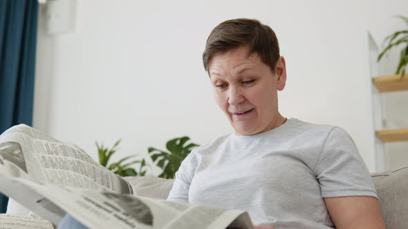 A Closeup View of a Beautiful Mature Woman is Reading a Newspaper Sitting on Sofa at Home in the