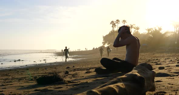 An attractive man with sunglasses relaxing on the beach at sunset as surfers walk along the shore in
